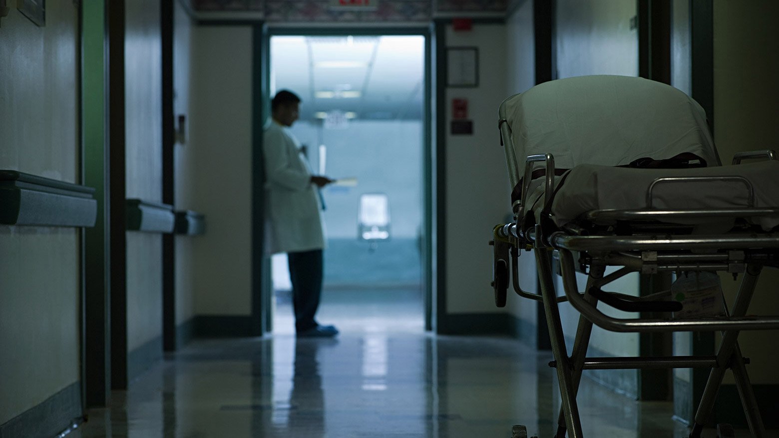 A doctor leans against a doorway in a hospital hallway containing a gurney