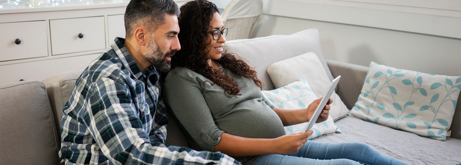 Pregnant woman and a man sitting on a couch looking at a tablet