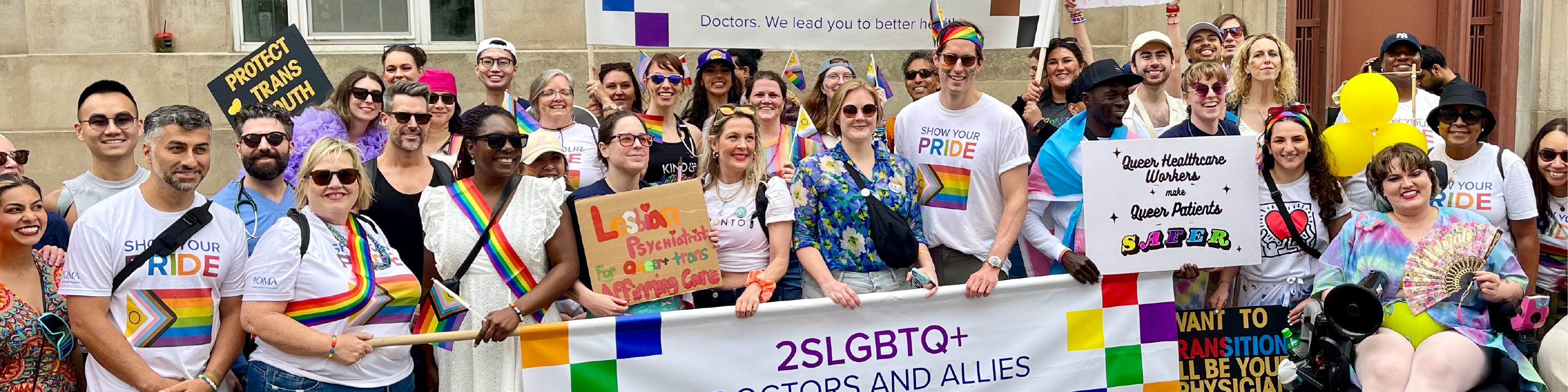 A large group of people wearing multicoloured clothing, with many holding signs, pose together with an OMA sign to celebrate Pride.