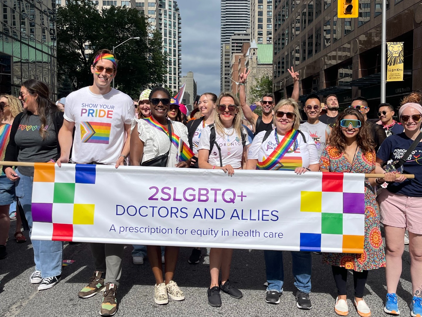 A group of people wearing Pride shirts stand in the middle of a street in downtown Toronto holding a sign that reads, “2SLGBTQ+ DOCTORS AND ALLIES, A prescription for equity in health care.”