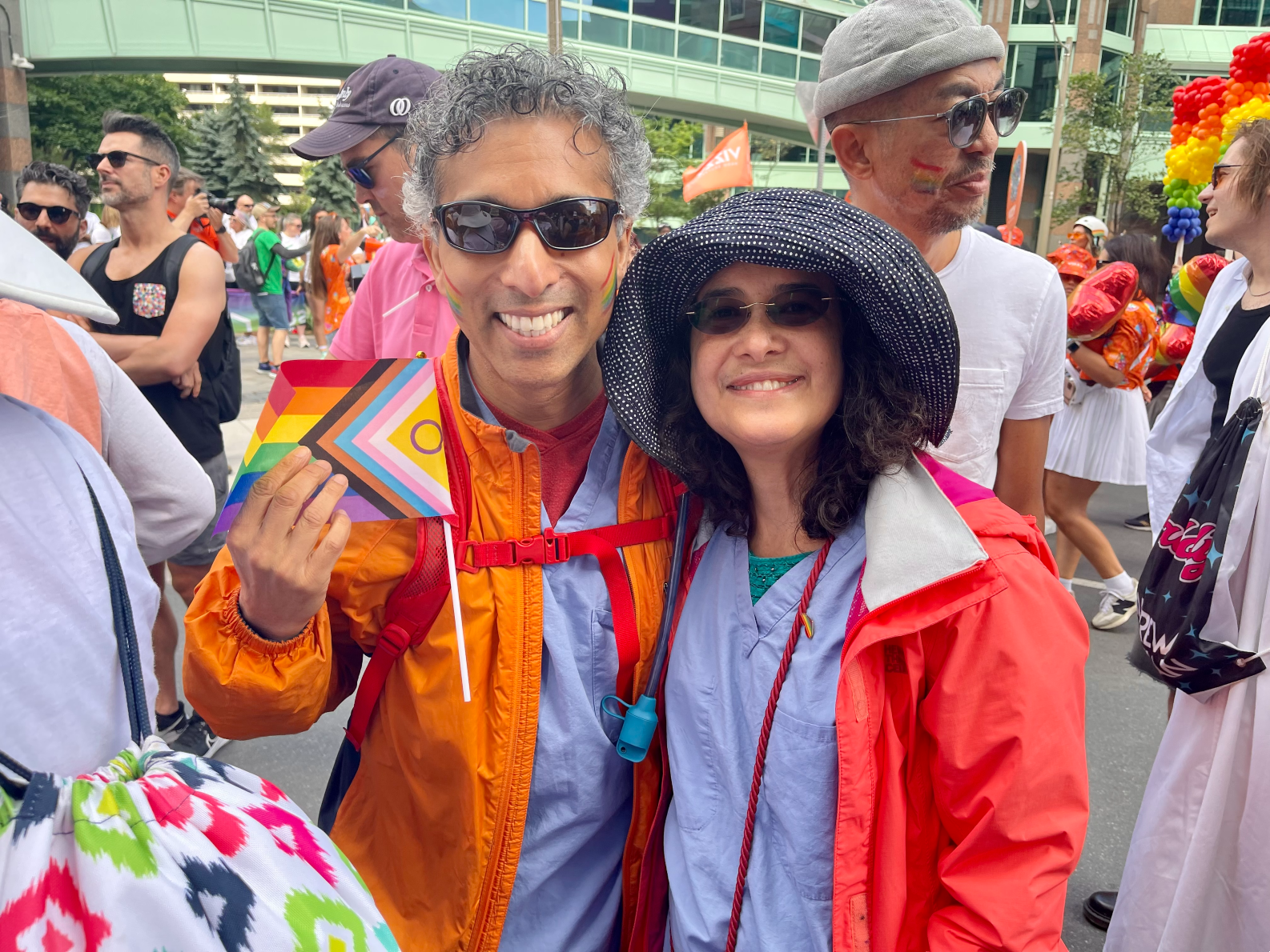 A man and woman wearing orange shirts and lavender scrubs smile while holding a Pride flag.