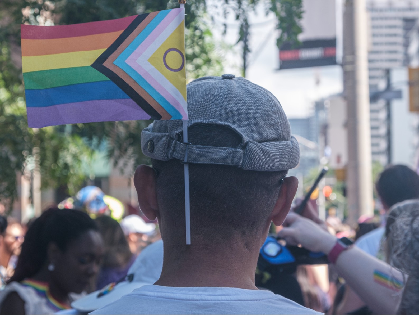 A picture of the back of the head of a man wearing a grey hat with a Pride flag stuck down the back.