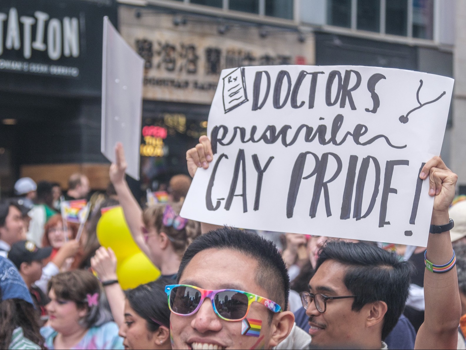A man wearing rainbow sunglasses with a Pride flag tattooed on his cheek smiles while a man behind him holds up a sign reading, ”DOCTORS prescribe GAY PRIDE!”