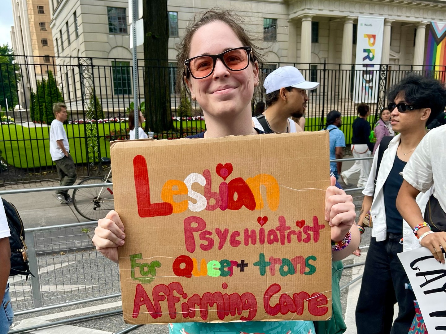 A woman wearing a long light blue dress and pink-tinted sunglasses smiles and holds a sign that reads, “Lesbian psychiatrist for Queer + trans Affirming Care.”